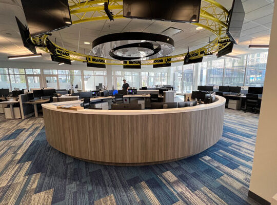 Modern office workspace featuring a curved wooden reception desk, surrounded by computer workstations and large screens, emphasizing custom woodworking and innovative design solutions.