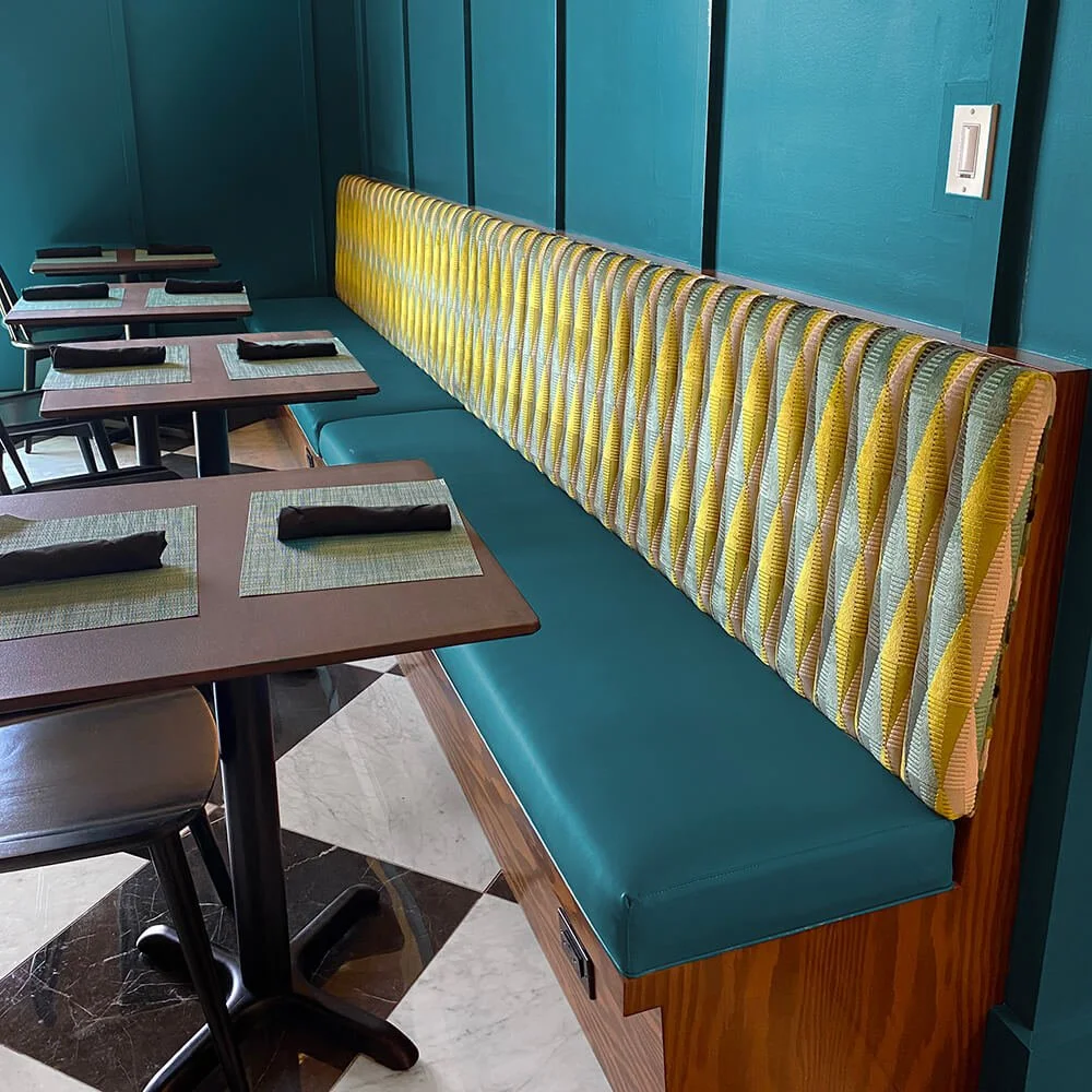 Stylish restaurant seating featuring a textured yellow and green upholstered bench against a teal wall, complemented by modern tables and black napkins, showcasing high craftsmanship and luxurious finishes.