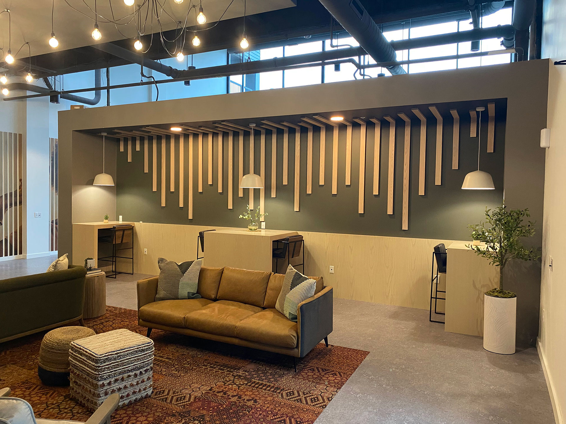 Modern office interior featuring a cozy seating area with a brown leather sofa, decorative pillows, and a patterned rug; minimalist workstations with wooden accents and pendant lighting; greenery and stylish decor elements enhancing the inviting atmosphere.