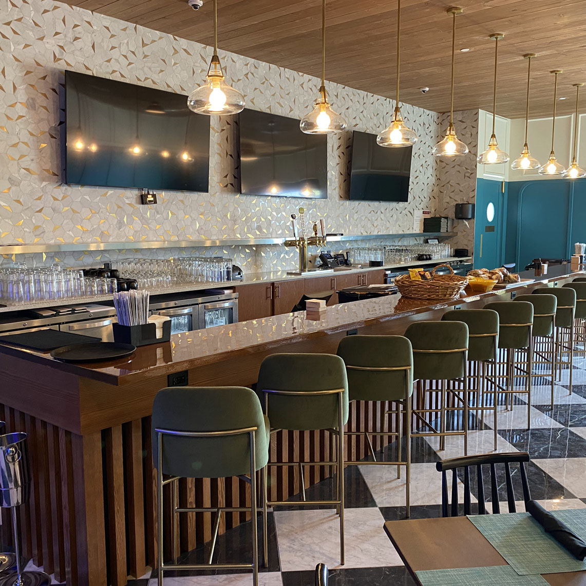 Modern bar interior featuring a sleek wooden counter, green upholstered stools, decorative wall with geometric patterns, and multiple flat-screen TVs, emphasizing quality craftsmanship and design.